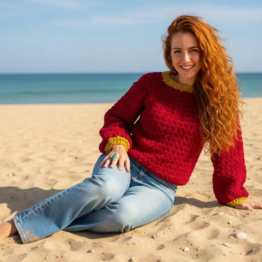 Pull au crochet fait main Pettirosso de Cachalabibi - rouge bordeaux - vue de face d'une jeune femme assise sur le sable d'une plage