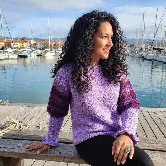 Pull au crochet fait main Fuga - violet lavande - vue de profil assise sur un banc à la marina avec voiliers