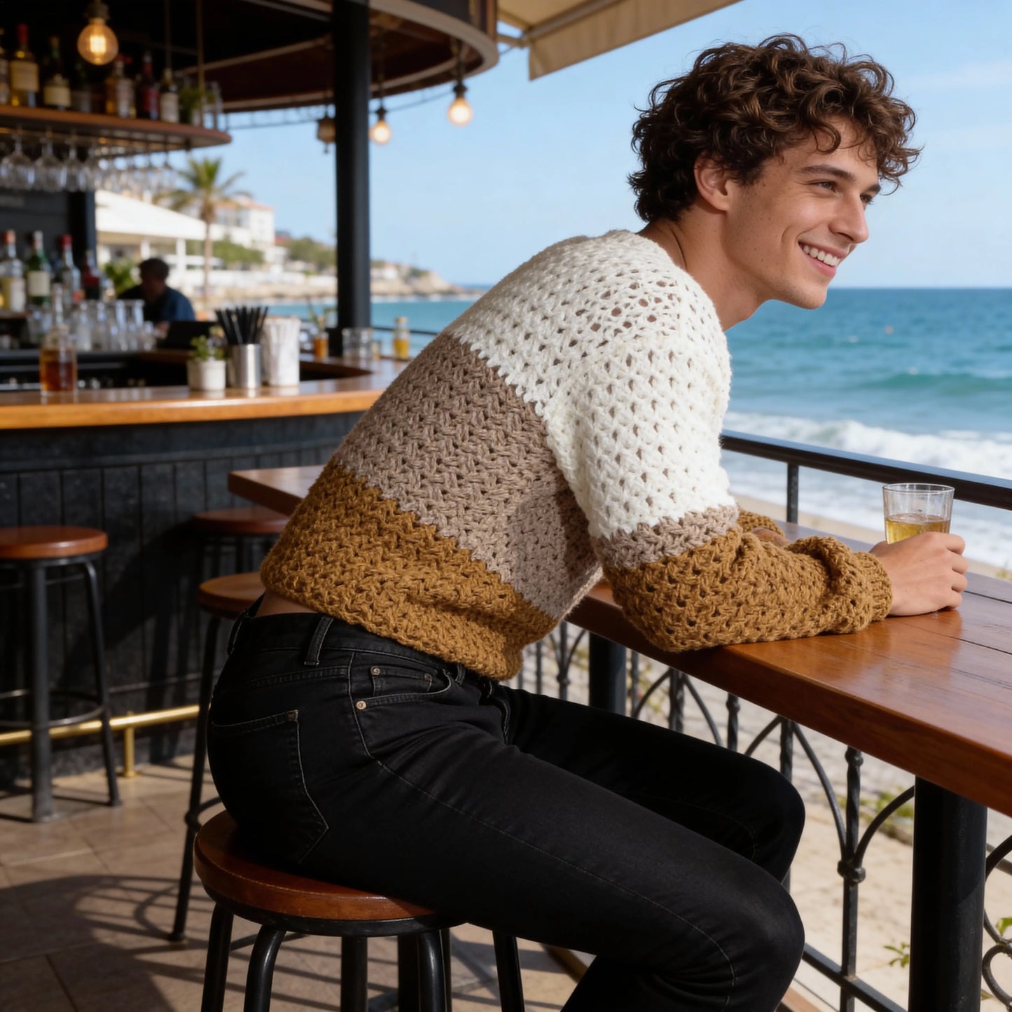 Pull au crochet fait main Homme Tricoloro de Cachalabibi - beige écru - vue de profil à la terrasse d'un bar de plage avec vue sur l'océan et verre de bière